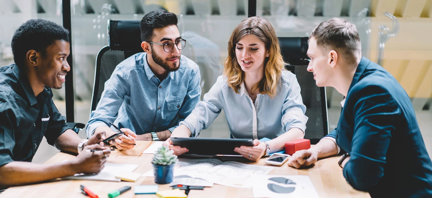 Een stockfoto waar drie mannen en één vrouw aan tafel zitten en met elkaar in gesprek zijn. De vrouw heeft een tablet in haar handen. 
