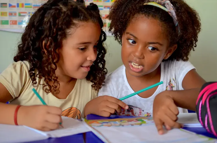 Een foto van twee kinderen in een klas, waarvan een kind een ander kind helpt met een opdracht