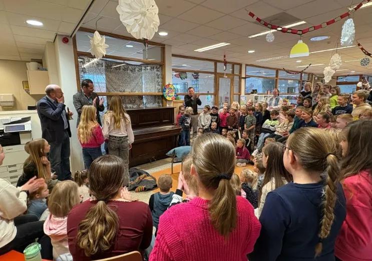 Groep met leerlingen om een piano heen met 3 volwassenen die klappen in hun handen.