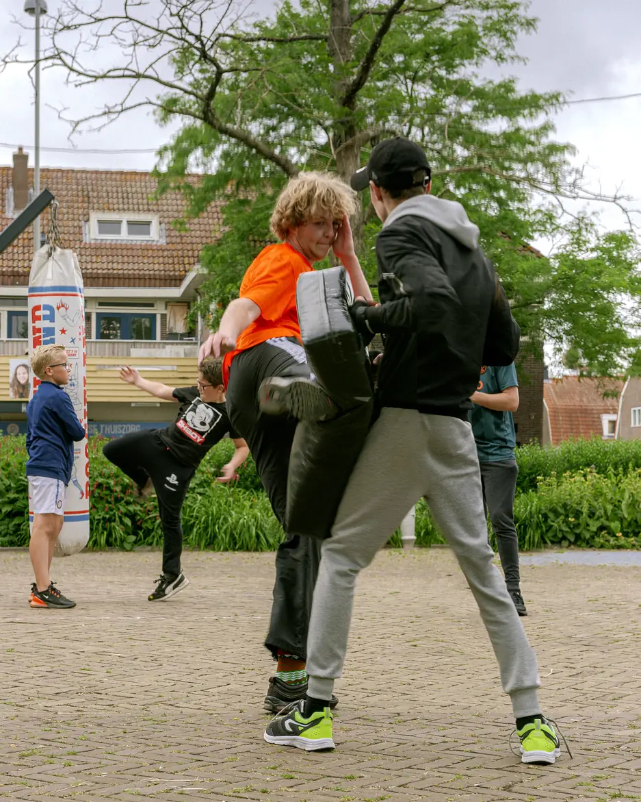 Jongeren oefenen kickbokstechnieken in de buitenlucht. Eén jongen in een oranje shirt trapt tegen een stootkussen dat wordt vastgehouden door een ander, terwijl op de achtergrond meer jongeren trainen.